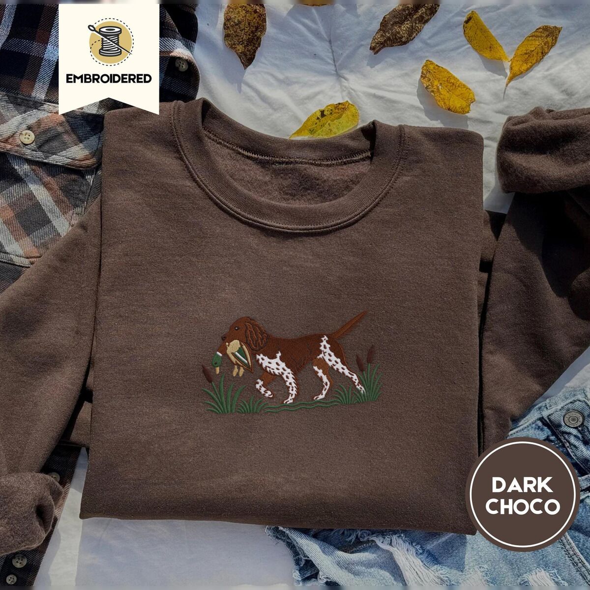 Bird Dog Embroidered Sweatshirt – Dark Chocolate Dark Chocolate crewneck sweatshirt on a plaid flannel shirt with yellow autumn leaves and denim shorts, embroidered with a liver and white bird dog carrying a mallard duck through cattails and marsh grass
