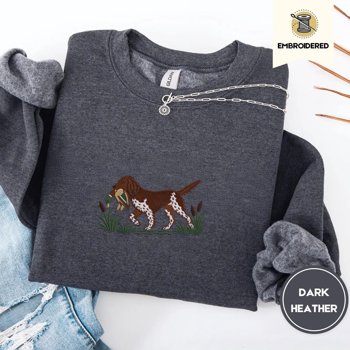 Bird Dog Embroidered Sweatshirt – Dark Heather Dark Heather crewneck sweatshirt folded with ripped jeans and a silver chain necklace, embroidered with a liver and white bird dog carrying a mallard duck through cattails and marsh grass
