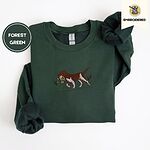 Forest Green crewneck sweatshirt folded on a white background, embroidered with a liver and white bird dog carrying a mallard duck through cattails and marsh grass