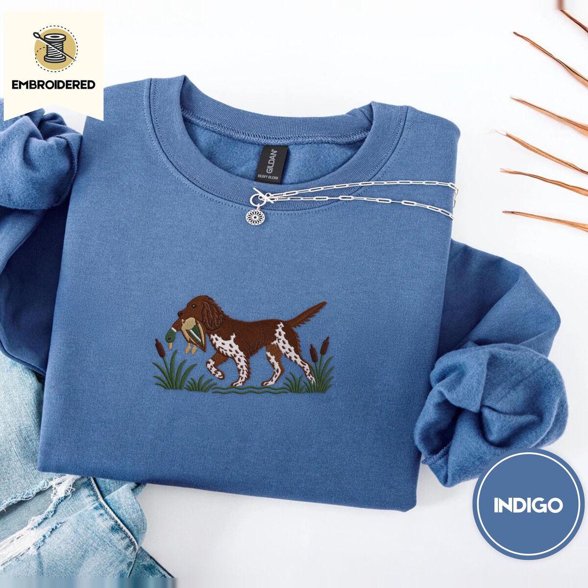 Bird Dog Embroidered Sweatshirt – Indigo Blue Indigo Blue crewneck sweatshirt folded with light jeans and dried twigs, embroidered with a liver and white bird dog carrying a mallard duck through cattails and marsh grass