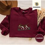 Maroon crewneck sweatshirt with a brown felt hat and dried bunny tail grass, embroidered with a liver and white bird dog carrying a mallard duck through cattails and marsh grass