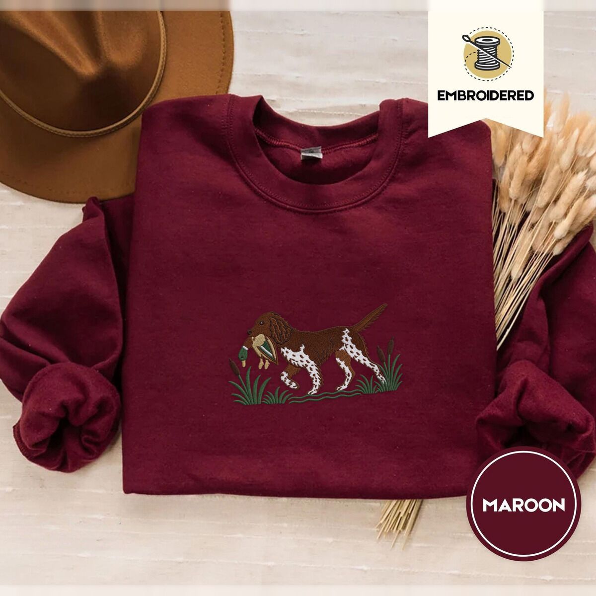 Bird Dog Embroidered Sweatshirt – Maroon Maroon crewneck sweatshirt with a brown felt hat and dried bunny tail grass, embroidered with a liver and white bird dog carrying a mallard duck through cattails and marsh grass