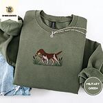 Military Green crewneck sweatshirt folded with light jeans and a silver chain necklace, embroidered with a liver and white bird dog carrying a mallard duck through cattails and marsh grass