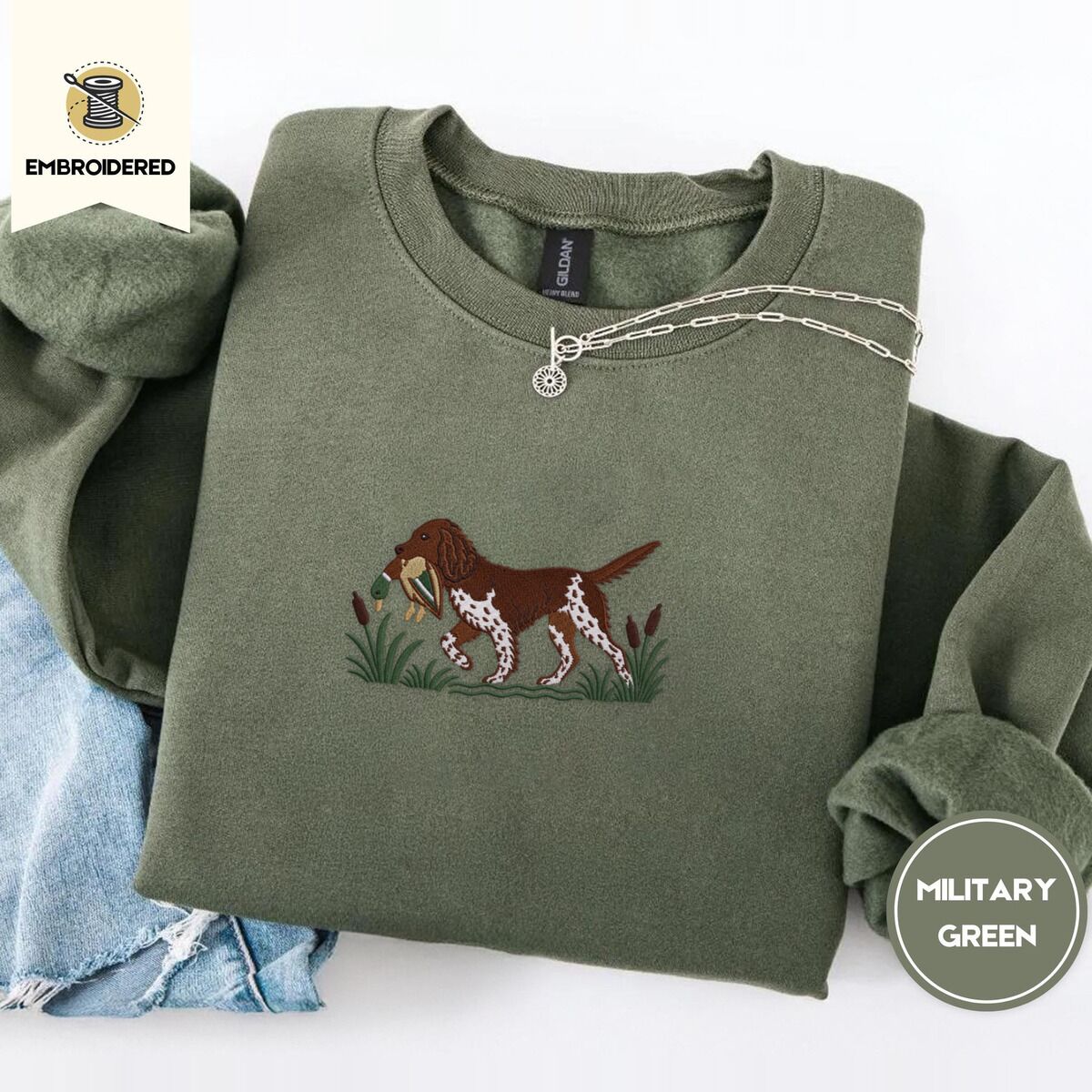 Bird Dog Embroidered Sweatshirt – Military Green Military Green crewneck sweatshirt folded with light jeans and a silver chain necklace, embroidered with a liver and white bird dog carrying a mallard duck through cattails and marsh grass
