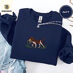 Navy crewneck sweatshirt folded with ripped jeans and a silver chain necklace, embroidered with a liver and white bird dog carrying a mallard duck through cattails and marsh grass