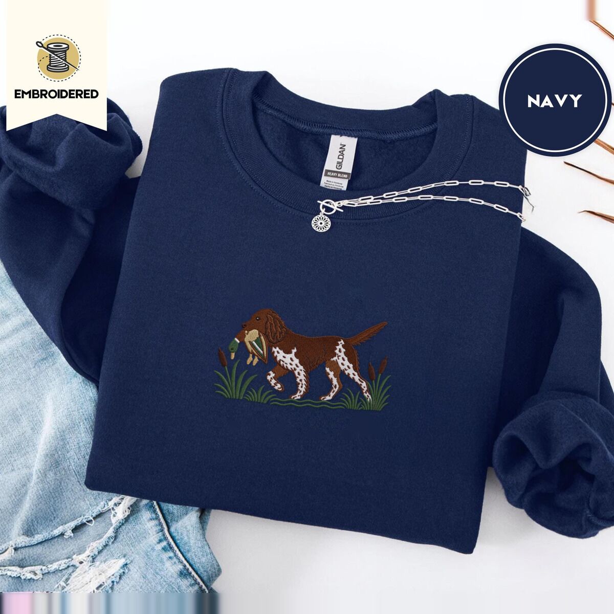 Bird Dog Embroidered Sweatshirt – Navy Navy crewneck sweatshirt folded with ripped jeans and a silver chain necklace, embroidered with a liver and white bird dog carrying a mallard duck through cattails and marsh grass
