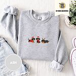 Ash crewneck sweatshirt folded with denim shorts, white sneakers and a potted plant, embroidered with three black and tan dachshunds in Santa hats, a red scarf and reindeer antlers across the chest