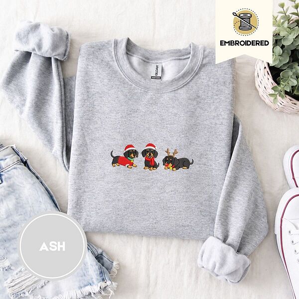 Ash crewneck sweatshirt folded with denim shorts, white sneakers and a potted plant, embroidered with three black and tan dachshunds in Santa hats, a red scarf and reindeer antlers across the chest