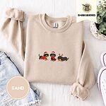 Sand crewneck sweatshirt folded with denim shorts, white sneakers and a potted plant, embroidered with three black and tan dachshunds in Santa hats, a red scarf and reindeer antlers across the chest