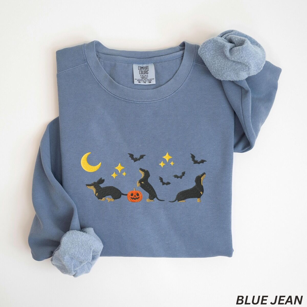 Black and Tan Dachshund Halloween Sweatshirt – Blue Jean Blue Jean crewneck sweatshirt laid flat on white, embroidered with three black and tan dachshunds playing under a yellow crescent moon, gold stars and flying bats beside a small jack-o'-lantern