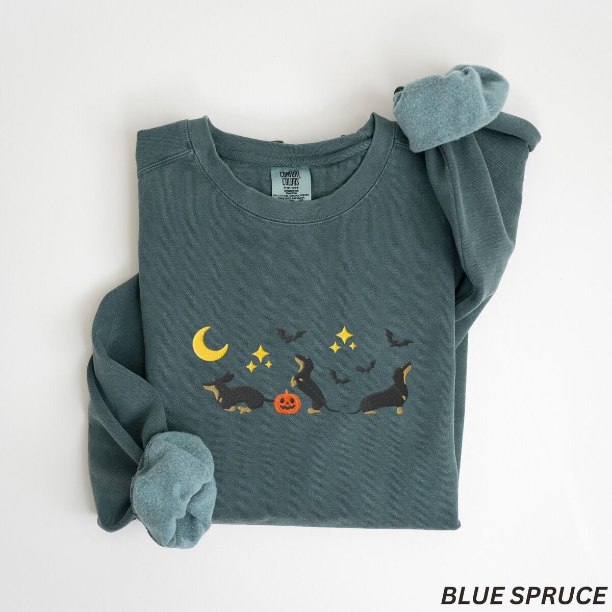 Black and Tan Dachshund Halloween Sweatshirt – Blue Spruce Blue Spruce crewneck sweatshirt laid flat on white, embroidered with three black and tan dachshunds playing under a yellow crescent moon, gold stars and flying bats beside a small jack-o'-lantern