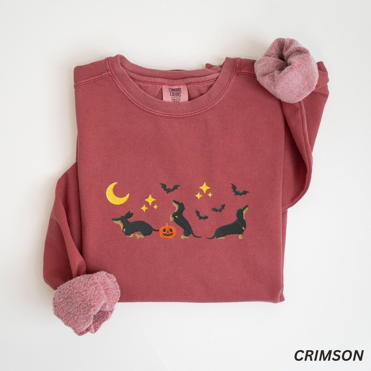Black and Tan Dachshund Halloween Sweatshirt – Crimson Crimson crewneck sweatshirt laid flat on white, embroidered with three black and tan dachshunds playing under a yellow crescent moon, gold stars and flying bats beside a small jack-o'-lantern