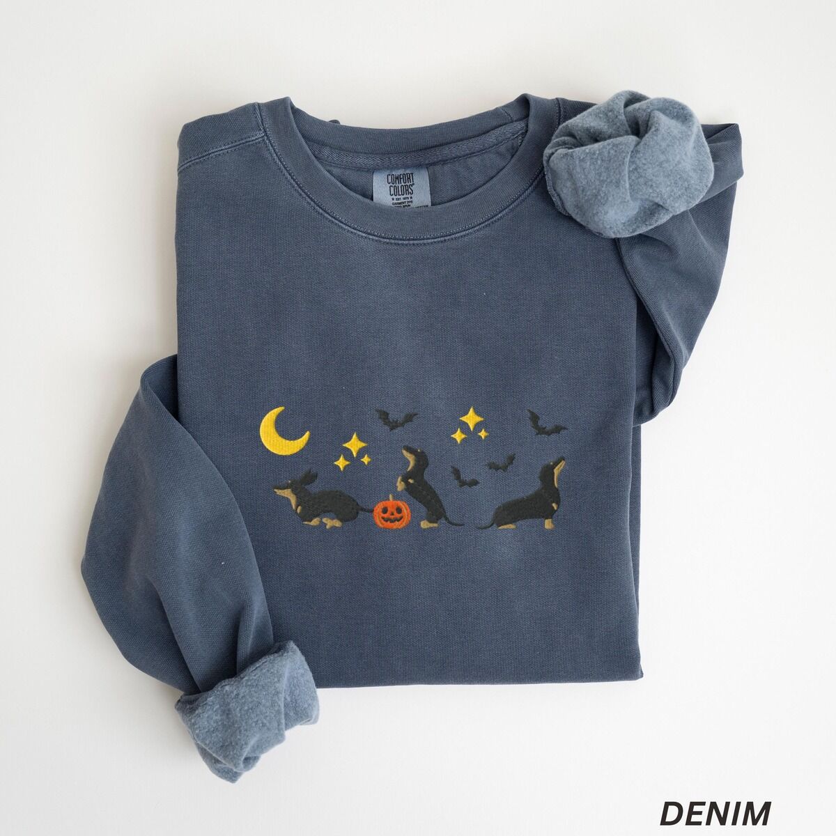 Black and Tan Dachshund Halloween Sweatshirt – Denim Denim crewneck sweatshirt laid flat on white, embroidered with three black and tan dachshunds playing under a yellow crescent moon, gold stars and flying bats beside a small jack-o'-lantern