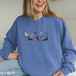 Smiling woman in a Flo Blue crewneck sweatshirt embroidered with three black and tan dachshunds playing under a yellow crescent moon, gold stars and flying bats beside a small jack-o'-lantern