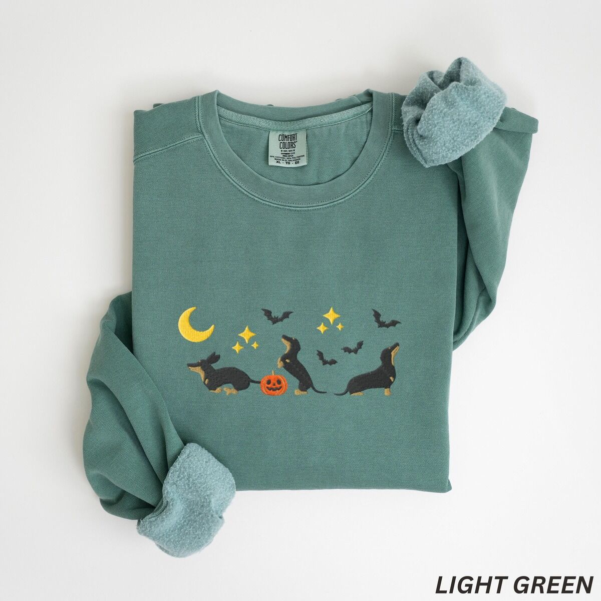 Black and Tan Dachshund Halloween Sweatshirt – Light Green Light Green crewneck sweatshirt laid flat on white, embroidered with three black and tan dachshunds playing under a yellow crescent moon, gold stars and flying bats beside a small jack-o'-lantern
