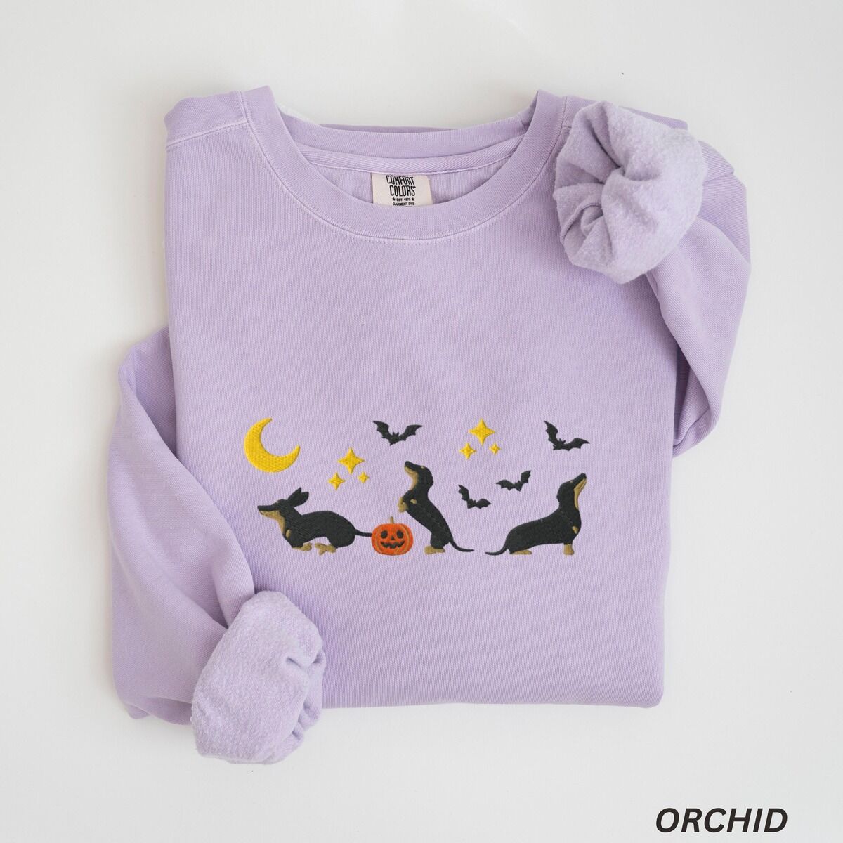 Black and Tan Dachshund Halloween Sweatshirt – Orchid Orchid crewneck sweatshirt laid flat on white, embroidered with three black and tan dachshunds playing under a yellow crescent moon, gold stars and flying bats beside a small jack-o'-lantern