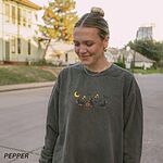Woman on a tree-lined street in a Pepper crewneck sweatshirt embroidered with three black and tan dachshunds playing under a yellow crescent moon, gold stars and flying bats beside a small jack-o'-lantern