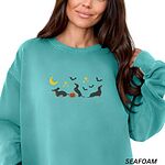 Woman in a Seafoam crewneck sweatshirt embroidered with three black and tan dachshunds playing under a yellow crescent moon, gold stars and flying bats beside a small jack-o'-lantern