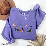 Violet crewneck sweatshirt laid flat with a hat and dried palm leaf, embroidered with three black and tan dachshunds playing under a yellow crescent moon, gold stars and flying bats beside a small jack-o'-lantern