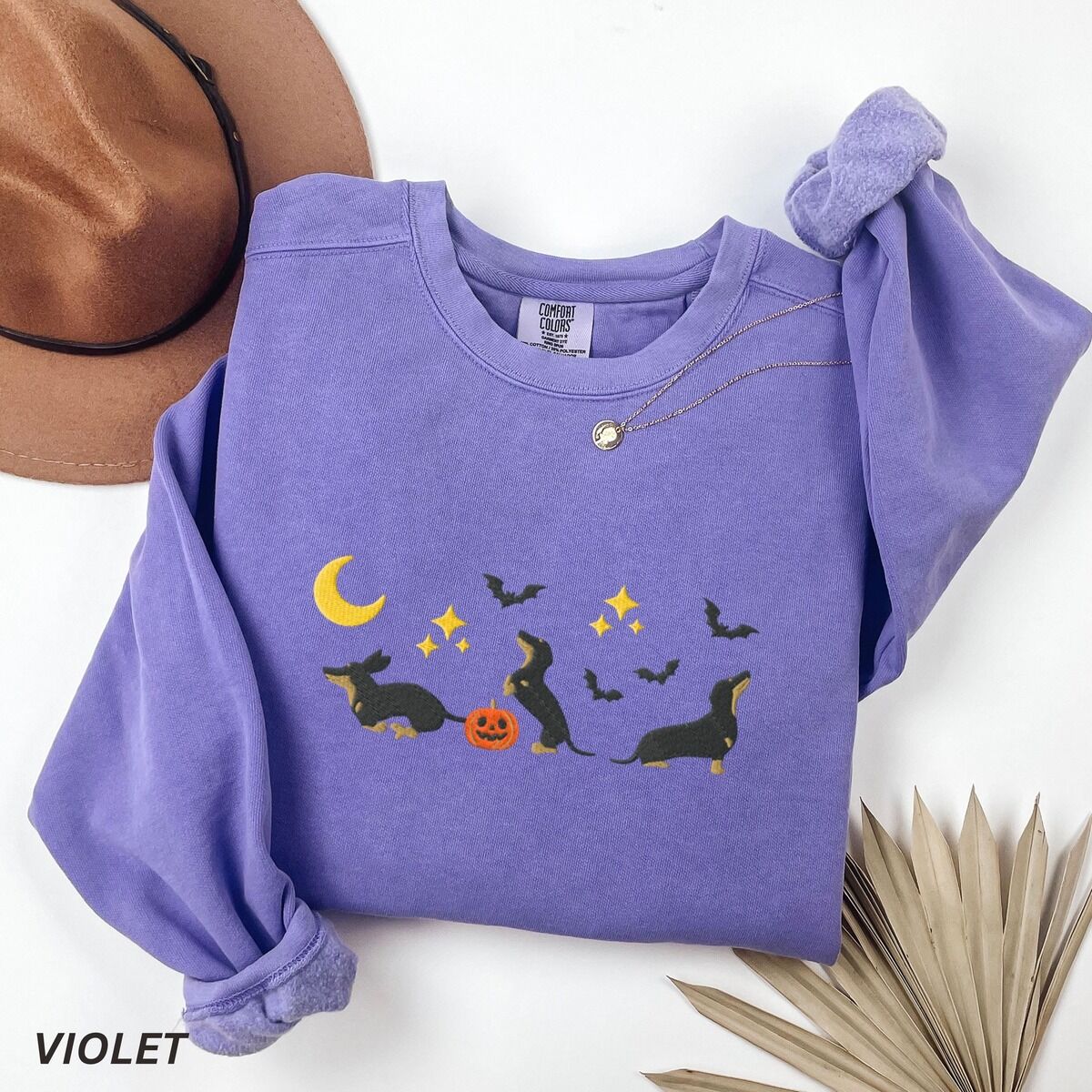 Black and Tan Dachshund Halloween Sweatshirt – Violet Violet crewneck sweatshirt laid flat with a hat and dried palm leaf, embroidered with three black and tan dachshunds playing under a yellow crescent moon, gold stars and flying bats beside a small jack-o'-lantern