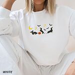 Woman sitting in a White crewneck sweatshirt embroidered with three black and tan dachshunds playing under a yellow crescent moon, gold stars and flying bats beside a small jack-o'-lantern