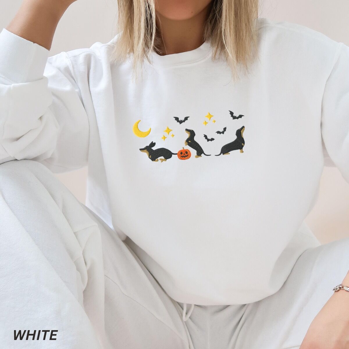 Black and Tan Dachshund Halloween Sweatshirt – White Woman sitting in a White crewneck sweatshirt embroidered with three black and tan dachshunds playing under a yellow crescent moon, gold stars and flying bats beside a small jack-o'-lantern