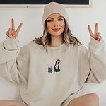 Woman in a beanie making peace signs in a Sand crewneck sweatshirt embroidered with a black and white pitbull in a Santa hat holding a candy cane beside a green polka dot gift box