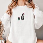 Woman in a White crewneck sweatshirt embroidered with a black and white pitbull in a Santa hat holding a candy cane beside a green polka dot gift box