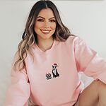 Smiling woman sitting in a Light Pink crewneck sweatshirt embroidered with a black and white pitbull in a Santa hat holding a candy cane beside a green polka dot gift box