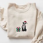 Sand crewneck sweatshirt laid flat, embroidered with a black and white pitbull in a Santa hat holding a candy cane beside a green polka dot gift box