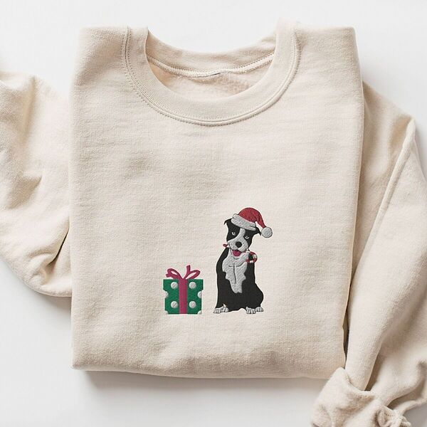 Sand crewneck sweatshirt laid flat, embroidered with a black and white pitbull in a Santa hat holding a candy cane beside a green polka dot gift box