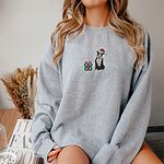 Woman sitting in an oversized Sport Grey crewneck sweatshirt embroidered with a black and white pitbull in a Santa hat holding a candy cane beside a green polka dot gift box