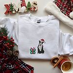 White crewneck sweatshirt laid flat with a plaid scarf, pinecones and a Santa hat, embroidered with a black and white pitbull in a Santa hat holding a candy cane beside a green polka dot gift box