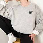Person in a light grey crewneck sweatshirt and black joggers, embroidered with a small minimalist black and white pit bull face with a white blaze and grey ears on the left chest