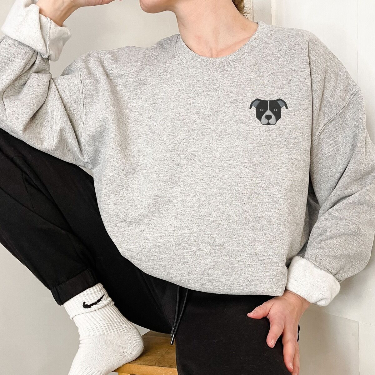 Black and White Pitbull Face Sweatshirt - worn in grey Person in a light grey crewneck sweatshirt and black joggers, embroidered with a small minimalist black and white pit bull face with a white blaze and grey ears on the left chest