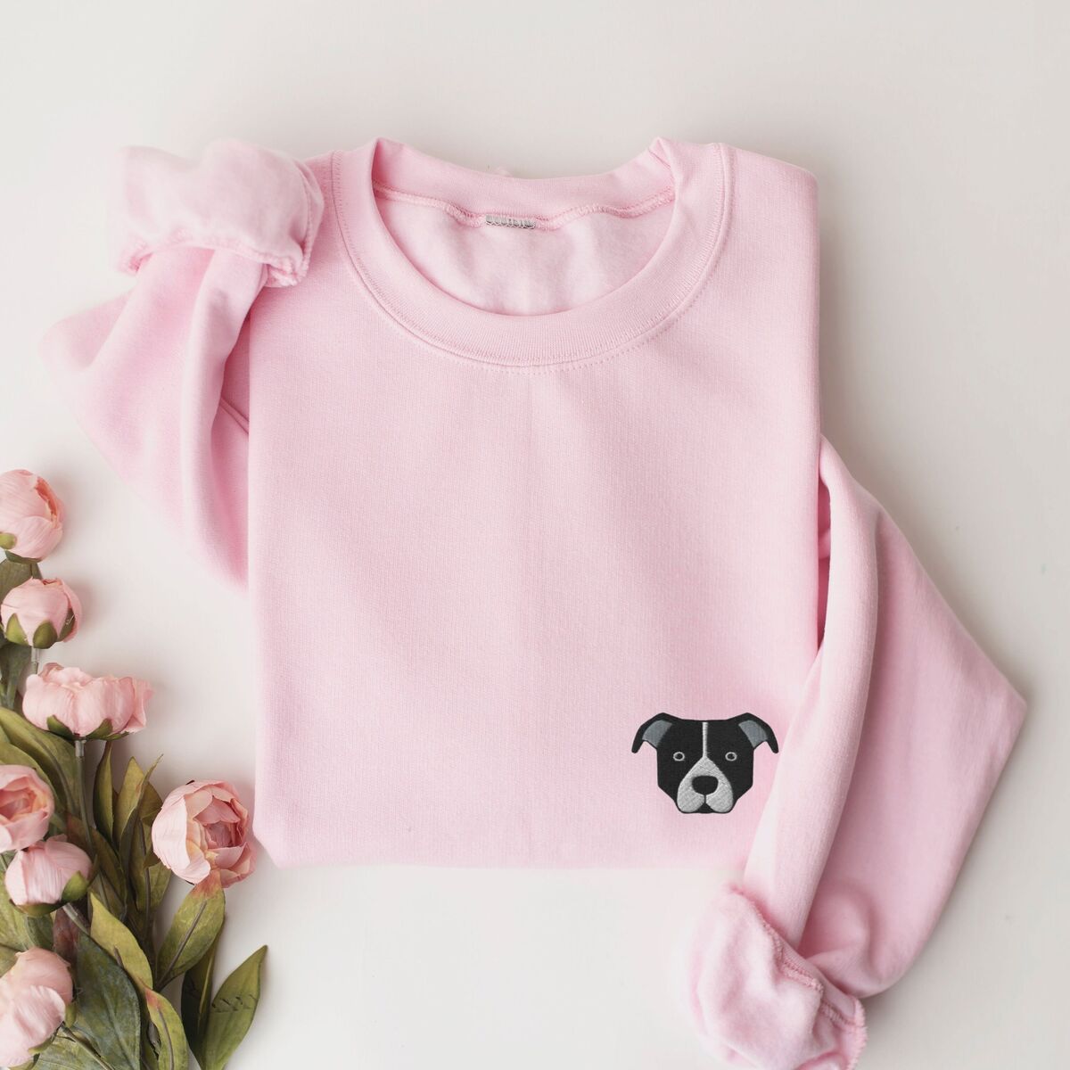 Black and White Pitbull Face Sweatshirt - Light Pink Light Pink crewneck sweatshirt laid flat with pink roses, embroidered with a small minimalist black and white pit bull face with a white blaze and grey ears on the left chest