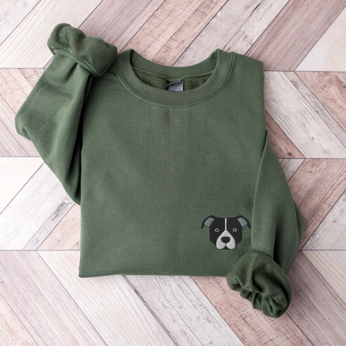 Black and White Pitbull Face Sweatshirt – Military Green Military Green crewneck sweatshirt laid flat on a wood floor, embroidered with a small minimalist black and white pit bull face with a white blaze and grey ears on the left chest