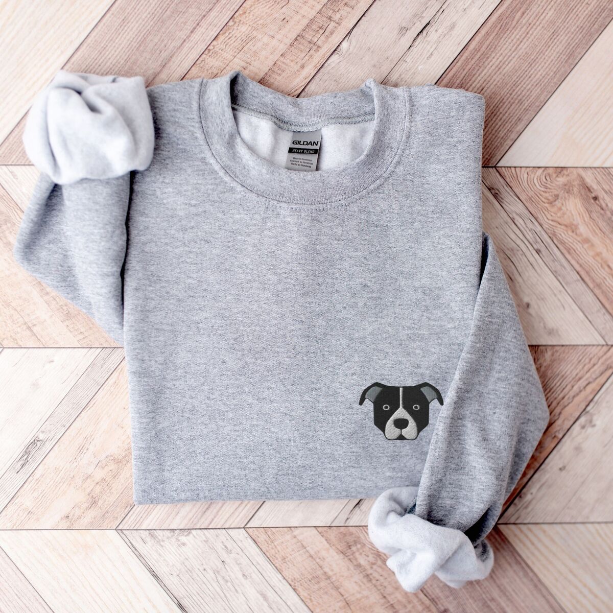 Black and White Pitbull Face Sweatshirt - Sport Grey Sport Grey crewneck sweatshirt laid flat on a wood floor, embroidered with a small minimalist black and white pit bull face with a white blaze and grey ears on the left chest