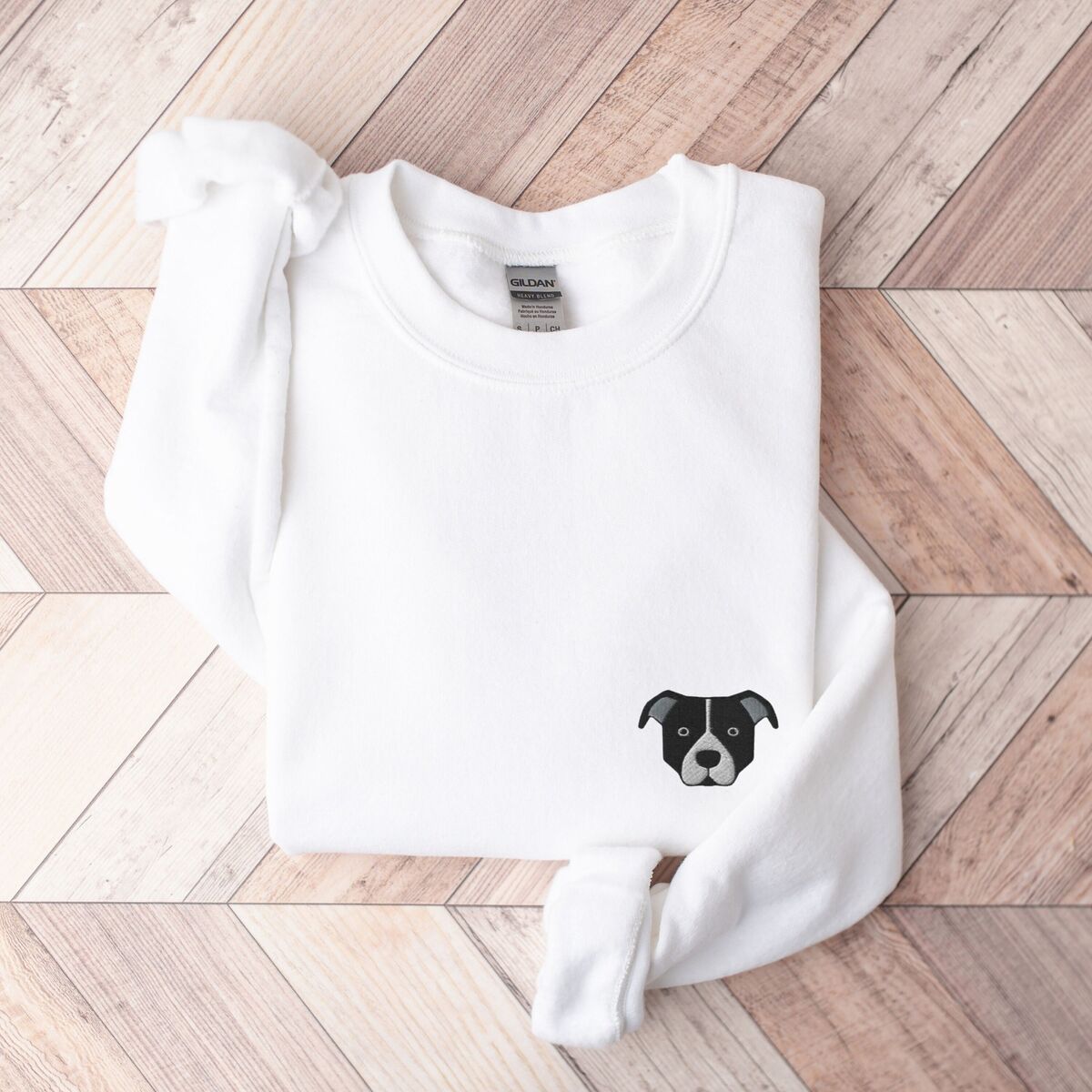 Black and White Pitbull Face Sweatshirt - White White crewneck sweatshirt laid flat on a wood floor, embroidered with a small minimalist black and white pit bull face with a white blaze and grey ears on the left chest
