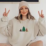 Woman in a knit beanie making peace signs in a Sand crewneck sweatshirt embroidered with a black lab in an elf hat and a yellow lab in a Santa hat and scarf beside a decorated Christmas tree with gifts