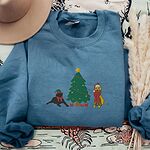 Indigo Blue crewneck sweatshirt laid flat with dried pampas, embroidered with a black lab in an elf hat and a yellow lab in a Santa hat and scarf beside a decorated Christmas tree with gifts