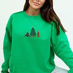 Woman sitting in an Irish Green crewneck sweatshirt embroidered with a black lab in an elf hat and a yellow lab in a Santa hat and scarf beside a decorated Christmas tree with gifts