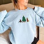 Woman in a Light Blue crewneck sweatshirt embroidered with a black lab in an elf hat and a yellow lab in a Santa hat and scarf beside a decorated Christmas tree with gifts