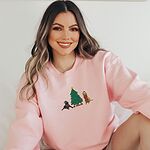 Smiling woman sitting in a Light Pink crewneck sweatshirt embroidered with a black lab in an elf hat and a yellow lab in a Santa hat and scarf beside a decorated Christmas tree with gifts