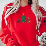 Woman by a Christmas tree in a Red crewneck sweatshirt embroidered with a black lab in an elf hat and a yellow lab in a Santa hat and scarf beside a decorated Christmas tree with gifts