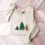 Sand crewneck sweatshirt laid flat on a chevron wood floor, embroidered with a black lab in an elf hat and a yellow lab in a Santa hat and scarf beside a decorated Christmas tree with gifts