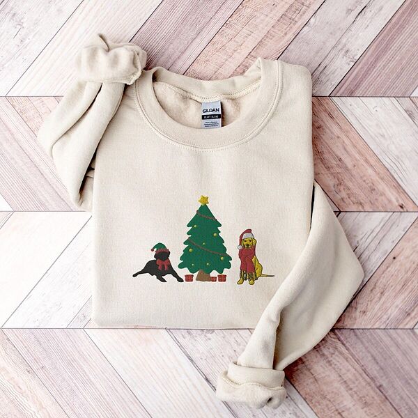 Sand crewneck sweatshirt laid flat on a chevron wood floor, embroidered with a black lab in an elf hat and a yellow lab in a Santa hat and scarf beside a decorated Christmas tree with gifts