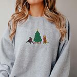 Woman in a Sport Grey crewneck sweatshirt embroidered with a black lab in an elf hat and a yellow lab in a Santa hat and scarf beside a decorated Christmas tree with gifts