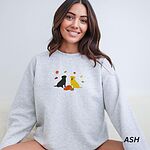 Smiling woman in an Ash crewneck sweatshirt embroidered with a black Labrador and a yellow Labrador sitting by three pumpkins under falling autumn leaves