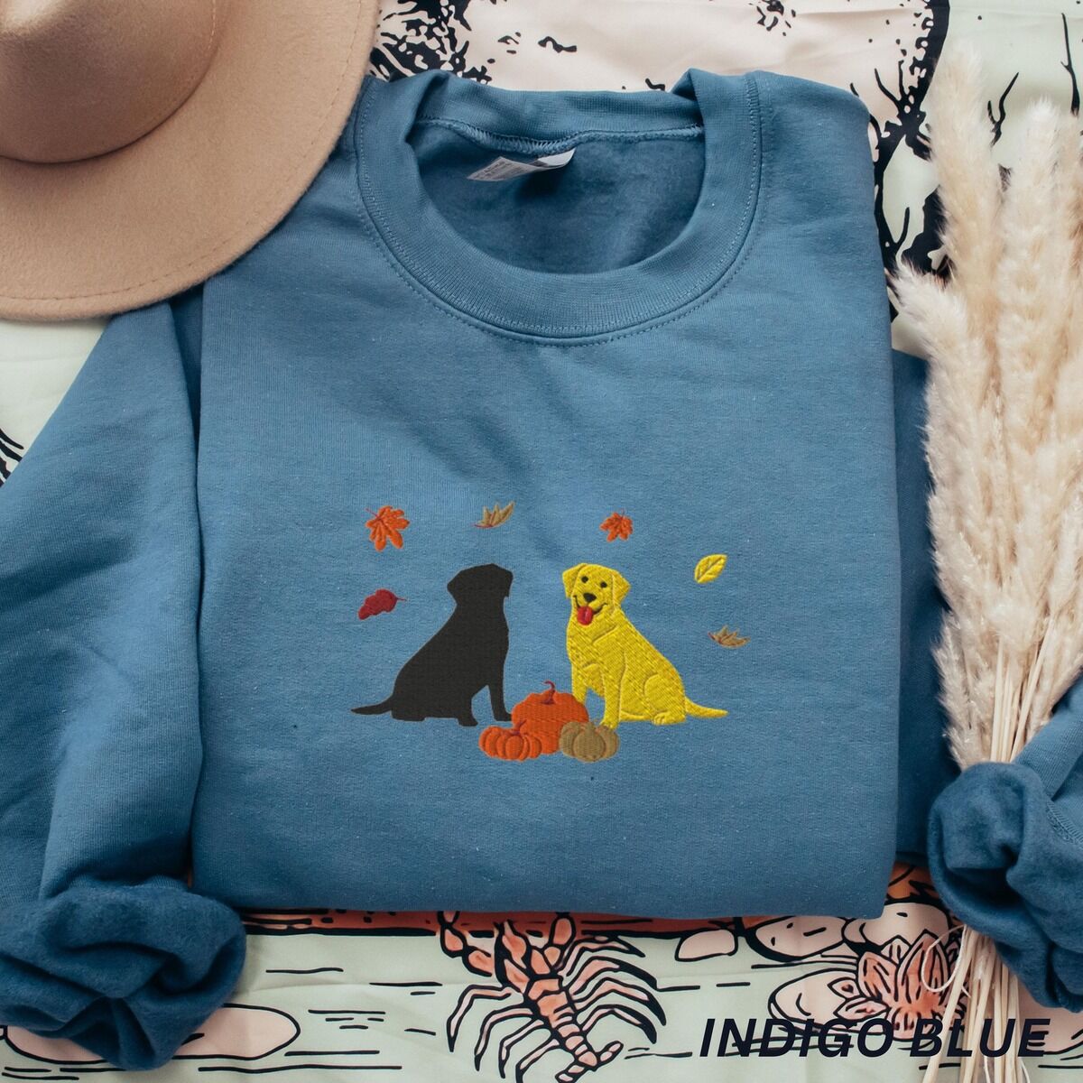 Black and Yellow Lab Fall Sweatshirt – Indigo Blue Indigo Blue crewneck sweatshirt laid flat with a hat and pampas grass, embroidered with a black Labrador and a yellow Labrador sitting by three pumpkins under falling autumn leaves